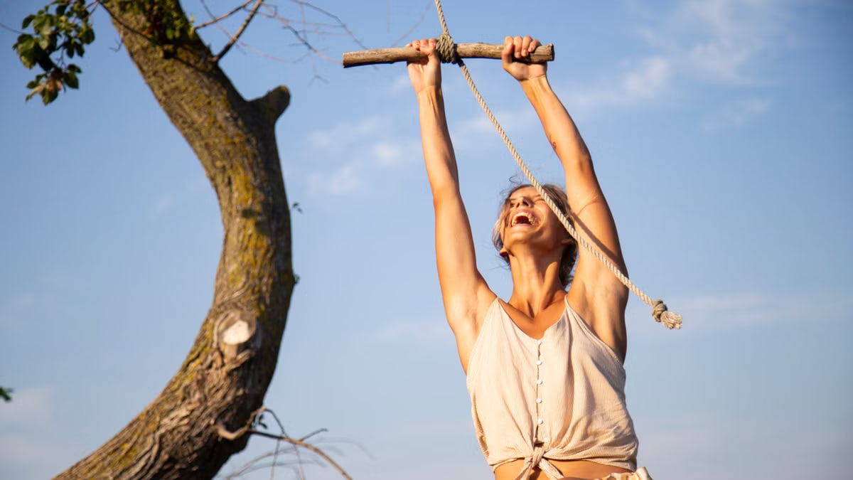 Happy woman enjoying an outdoor adventure, swinging on a wooden stick rope swing under a sunny sky.